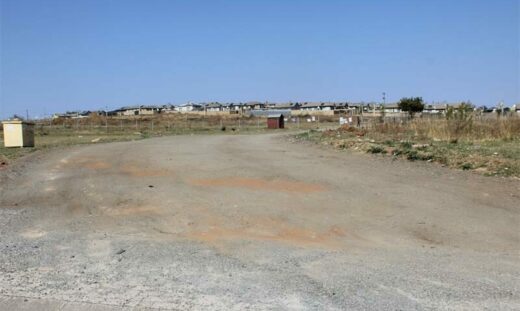 A vast, empty dirt road stretches under a clear blue sky. Sparse grass grows along the roadside, with a distant view of low-profile buildings. Quiet and desolate.