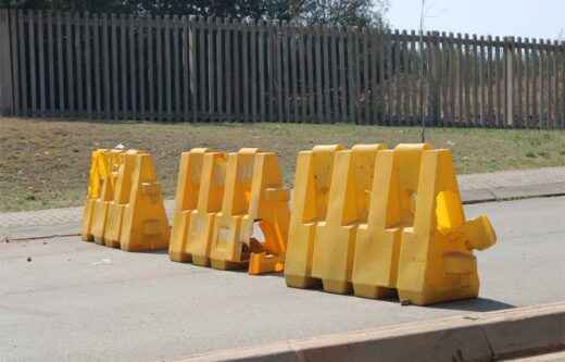 Yellow road barriers line a deserted street under clear skies, set against a wooden fence and dry grass, conveying a sense of quiet restriction.