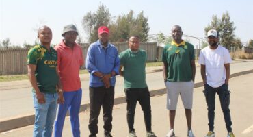 A group of six men standing together on a paved street, posing for a photo. They are casually dressed, with some wearing sports shirts and caps, and a wooden fence with trees is visible in the background.