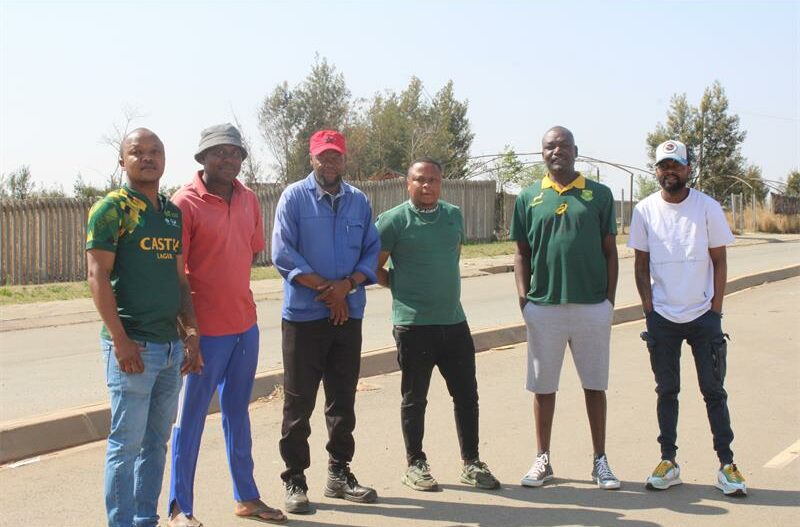 A group of six men standing together on a paved street, posing for a photo. They are casually dressed, with some wearing sports shirts and caps, and a wooden fence with trees is visible in the background.