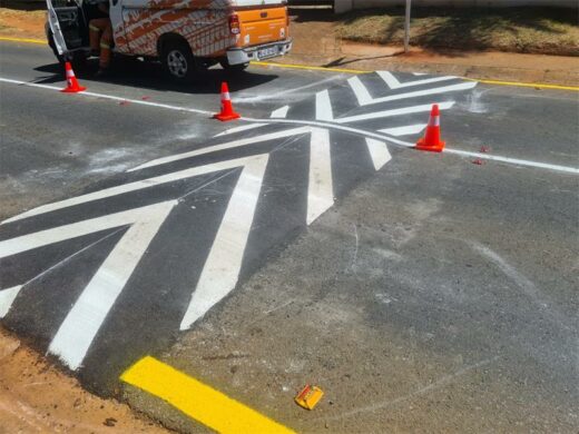 Road with optical illusion stripes resembling a zigzag pattern, surrounded by orange traffic cones. A vehicle is parked nearby, adding contrast.