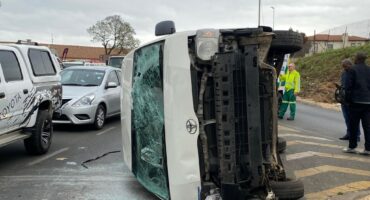 A white taxi lies on its side after a collision on a road, with shattered glass nearby. Emergency responders and bystanders are present at the scene.