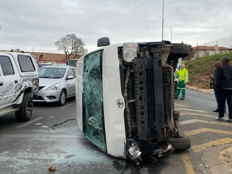 A white taxi lies on its side after a collision on a road, with shattered glass nearby. Emergency responders and bystanders are present at the scene.