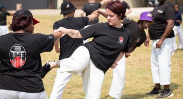 A martial arts class practices outdoors on grass. Participants in black shirts and white pants engage in synchronized moves, showing focus and teamwork.