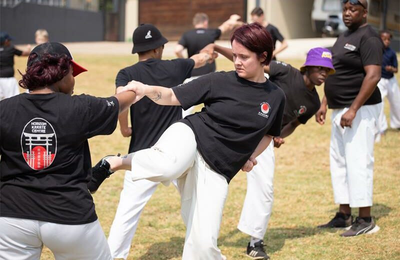 A martial arts class practices outdoors on grass. Participants in black shirts and white pants engage in synchronized moves, showing focus and teamwork.