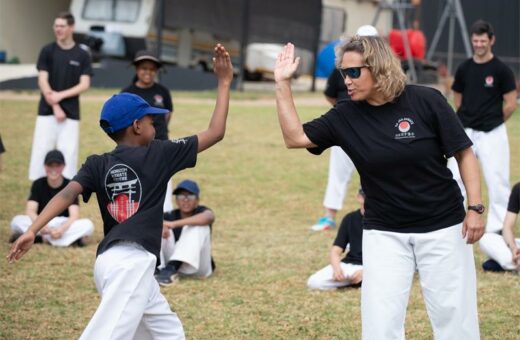 A young boy and an adult woman in martial arts uniforms exchange high-fives outdoors, showing teamwork and encouragement. Others sit and smile in the background.