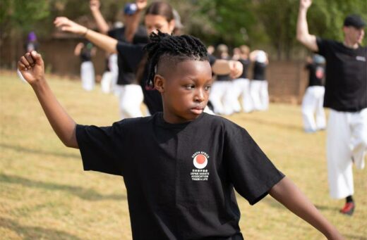 A young person practices martial arts outdoors, wearing a black T-shirt with a logo. Others in similar attire move in unison, exuding focus and coordination.