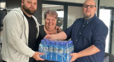 Three people hold a pack of bottled water indoors. They are smiling, conveying a sense of teamwork and camaraderie in a casual setting.