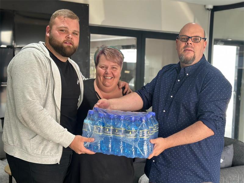 Three people hold a pack of bottled water indoors. They are smiling, conveying a sense of teamwork and camaraderie in a casual setting.