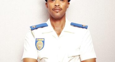 Man in a white police uniform with blue and gold accents stands confidently with arms crossed against a neutral background, conveying authority and professionalism.