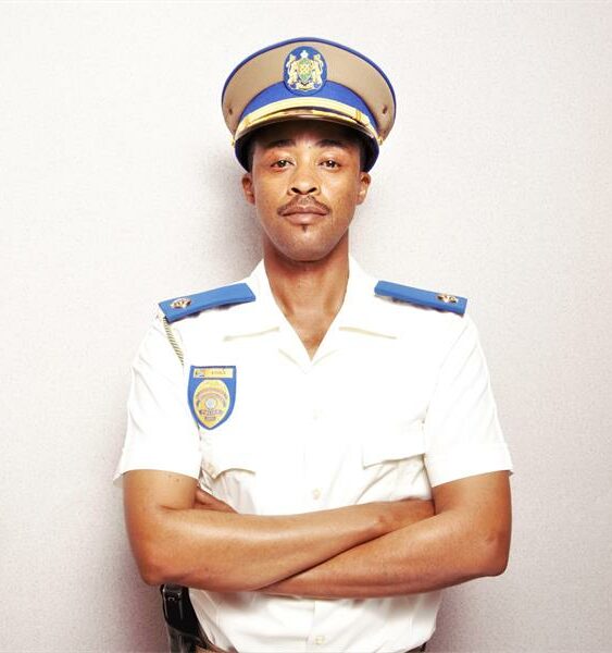 Man in a white police uniform with blue and gold accents stands confidently with arms crossed against a neutral background, conveying authority and professionalism.