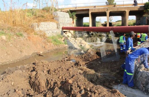 Workers in blue uniforms repair a riverbank near a bridge. A red pipe runs across, with rocks and wire mesh visible. The setting is industrious.