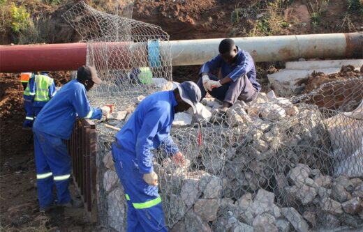 Two workers in blue uniforms and gloves build a stone gabion wall near a large pipe. One sits on rocks, focusing intently. The scene is industrious.