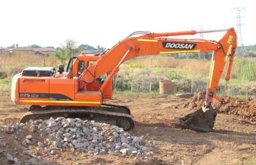 An orange excavator labeled "Doosan 225 LCV" is working on a construction site, its bucket digging into the dirt. Piles of rocks and green fields are in the background under a clear sky.