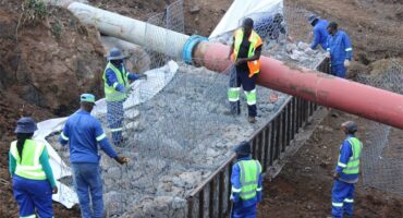 Construction workers in bright safety vests and helmets secure a large pipeline using wire mesh and rocks in a muddy, earth-filled trench. The scene is busy and industrious.