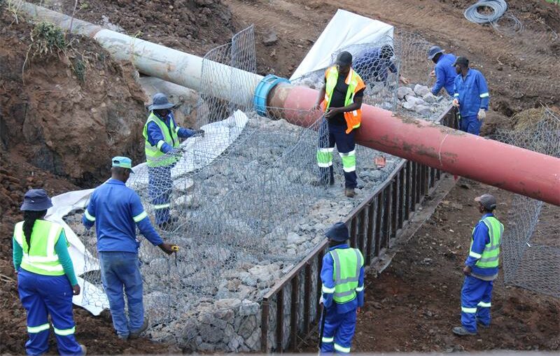 Construction workers in bright safety vests and helmets secure a large pipeline using wire mesh and rocks in a muddy, earth-filled trench. The scene is busy and industrious.
