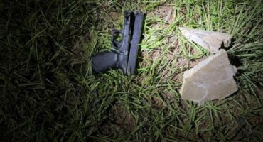 A black handgun lies on grass, illuminated by a flashlight. Two broken stones are nearby, creating a tense and mysterious atmosphere.