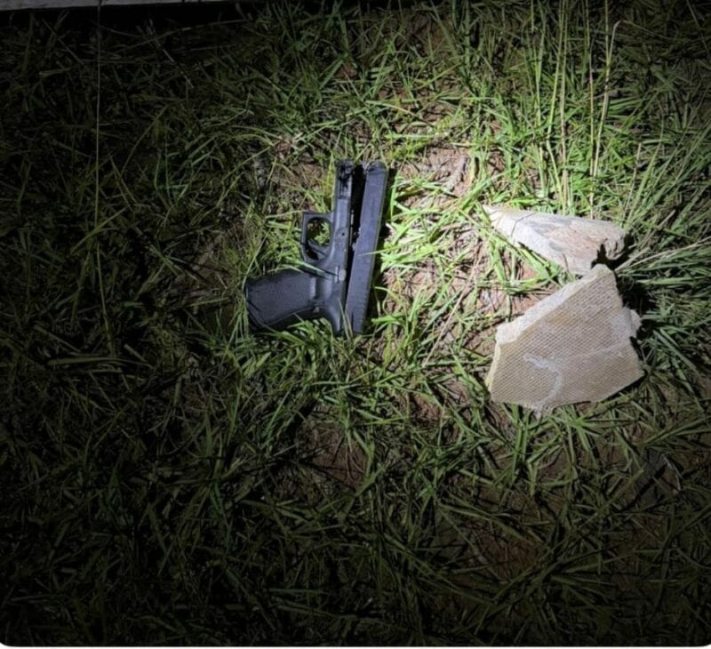 A black handgun lies on grass, illuminated by a flashlight. Two broken stones are nearby, creating a tense and mysterious atmosphere.