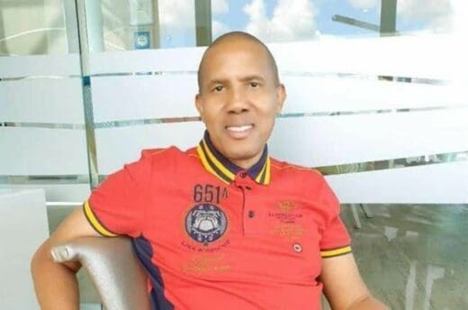 Man sitting in front of house, wearing a red shirt.