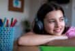 Young girl wearing headphones, smiling while sitting at a desk with colourful pencils.