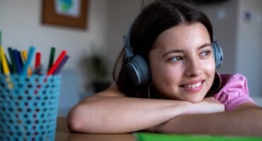 Young girl wearing headphones, smiling while sitting at a desk with colourful pencils.