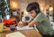 A young boy wearing headphones studies at a desk, writing in a notebook with a pencil beside a tablet.