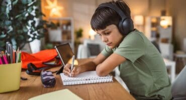 A young boy wearing headphones studies at a desk, writing in a notebook with a pencil beside a tablet.