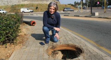 Tracy Kensey kneels beside an uncovered stormwater drain on the pedestrian walkway at the corner of Rivonia and Leeuwkop roads in Sunninghill as she raises concerns about the potential danger it poses to the public on 28 June, 2026. Photo: Ayanda Ntshingila