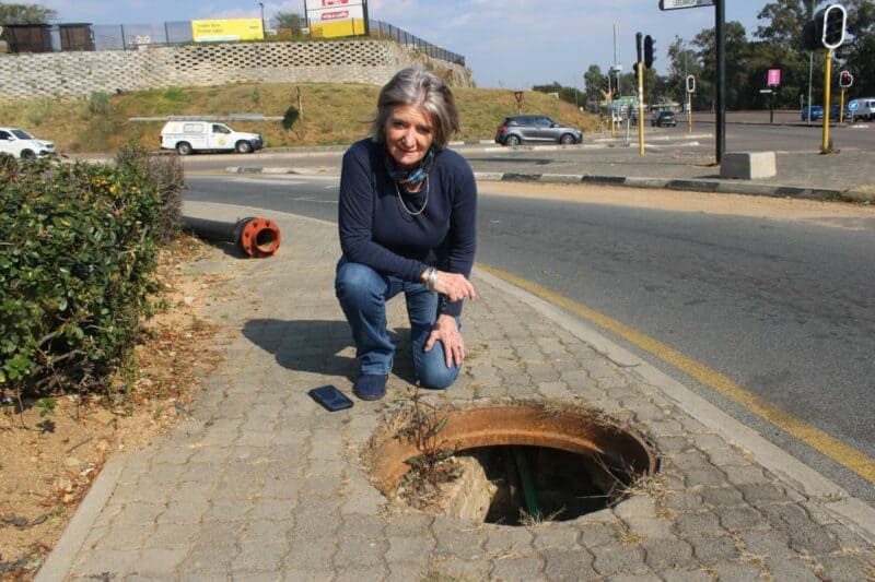 Tracy Kensey kneels beside an uncovered stormwater drain on the pedestrian walkway at the corner of Rivonia and Leeuwkop roads in Sunninghill as she raises concerns about the potential danger it poses to the public on 28 June, 2026. Photo: Ayanda Ntshingila