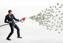 A smiling businessman holding a gas nozzle while many dollar bills are flying out of it instead of petrol.