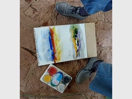 A sketch pad and paint pallette on the ground between a person's feet.