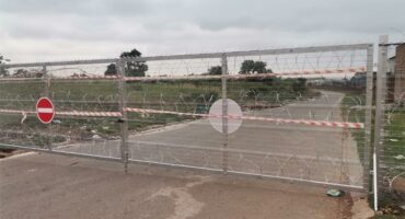 An access control gate installed on a public road, with barbed wire and barrier tape red and white.