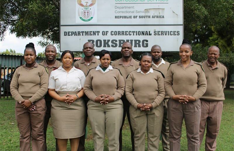 Boksburg Correctional Services officials, dressed in brown correctional uniforms, stand behind the Boksburg Correctional Services signage.