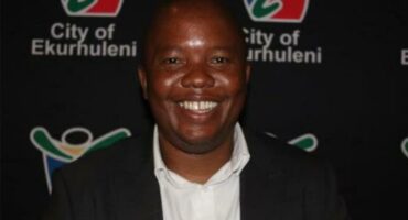 A black man in a black blazer jacket and a white shirt, standing with a black banner bearing the City of Ekurhuleni logo.