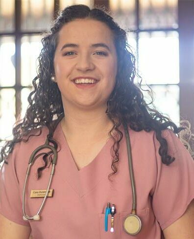 Woman with dark curly hair wearing pink scrubs sitting in front of a brightly lit window