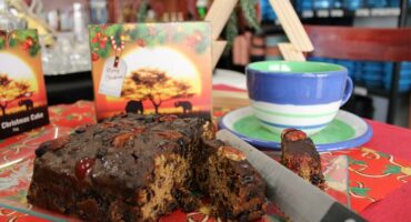 Bread knife cutting into a fruit cake with a cup and packaging in the background