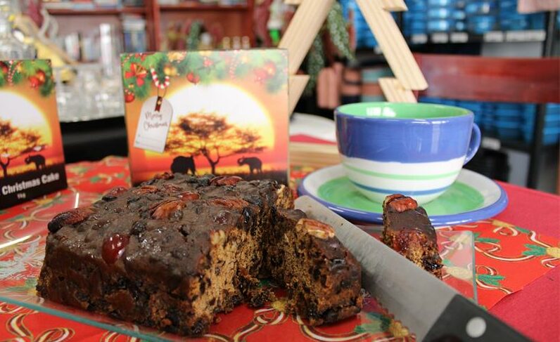 Bread knife cutting into a fruit cake with a cup and packaging in the background