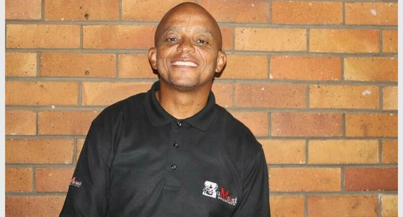 “Black man wearing a black top standing in front of a brown brick wall.