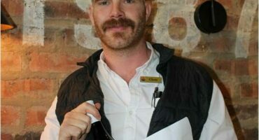 A man with mustache wearing a white shirt and black sleeveless jacket, and holding a bottle.