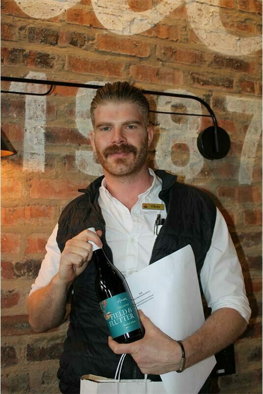A man with mustache wearing a white shirt and black sleeveless jacket, and holding a bottle.