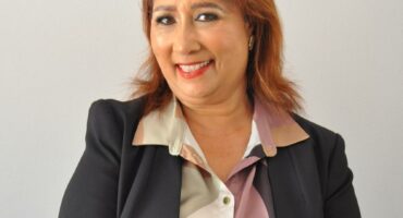 Woman with brown hair wearing a grey outfit, standing against a white background.