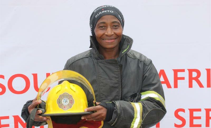 Black female firefighter in full protective gear holding her helmet.