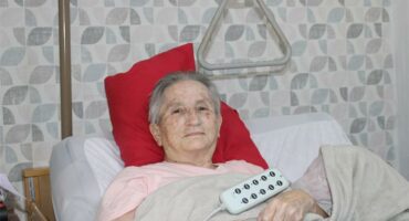 Grandmother lying in bed supported by a red pillow.