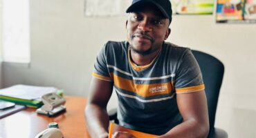 A Black man wearing a cap seated on a chair in an office