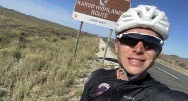 White cyclist wearing a white helmet standing with a bicycle in front of a sign that reads “Karoo Highlands Route."