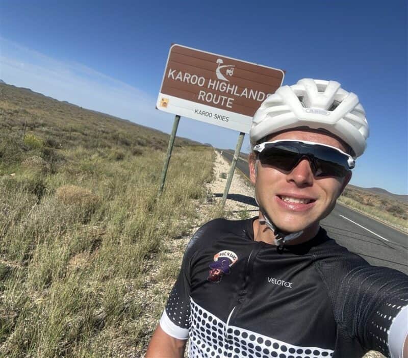 White cyclist wearing a white helmet standing with a bicycle in front of a sign that reads “Karoo Highlands Route.