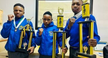 Three schoolboys wearing blue school jerseys pose for a photo while holding trophies.