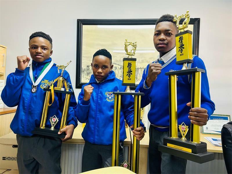 Three schoolboys wearing blue school jerseys pose for a photo while holding trophies.