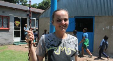 A white woman standing , with a school building and children in the background