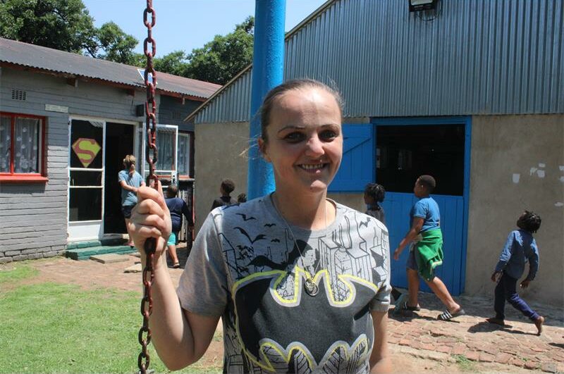 A white woman standing , with a school building and children in the background